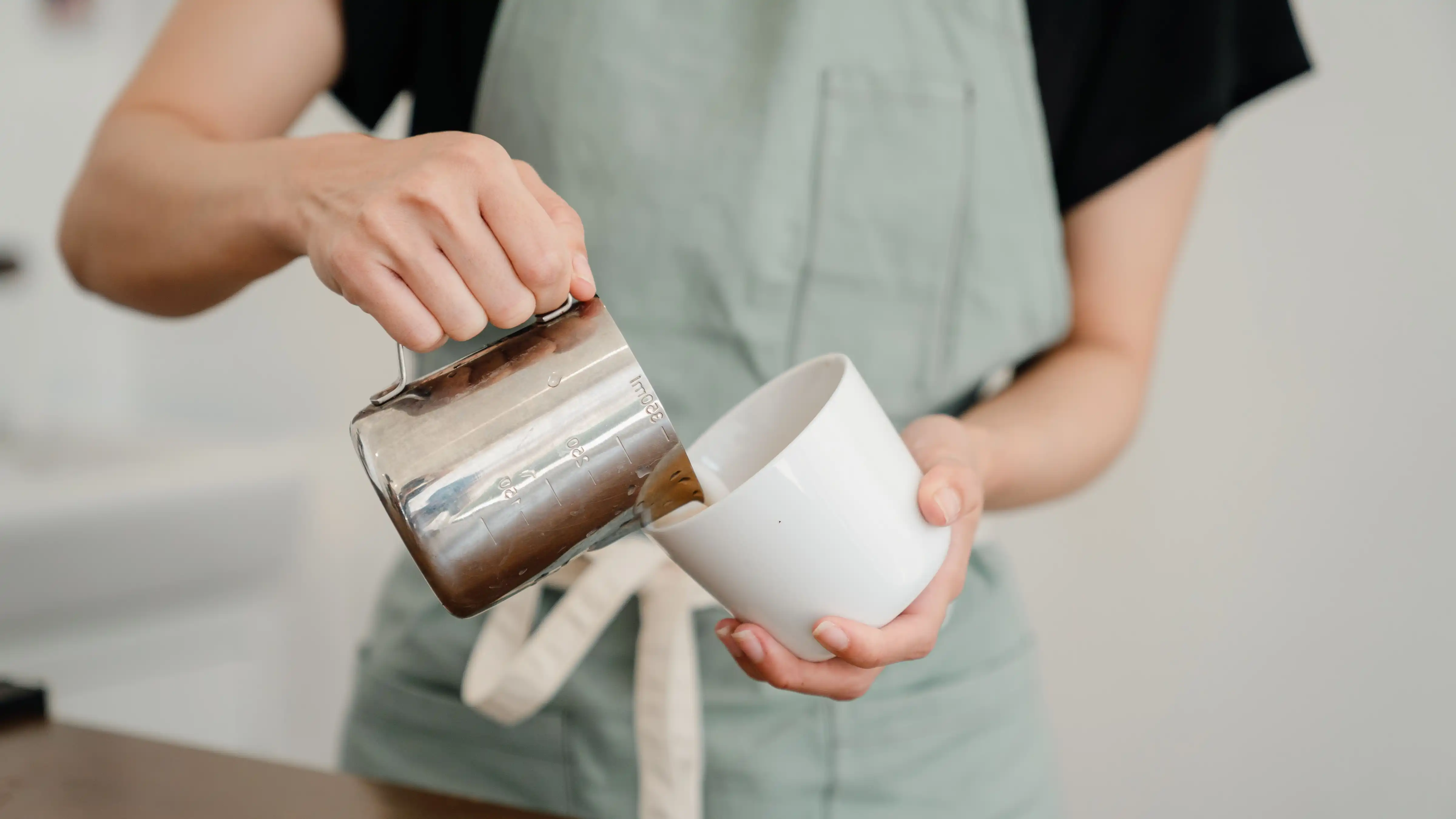 A barista in an apron pouring frothed milk from a stainless steel pitcher into a white mug